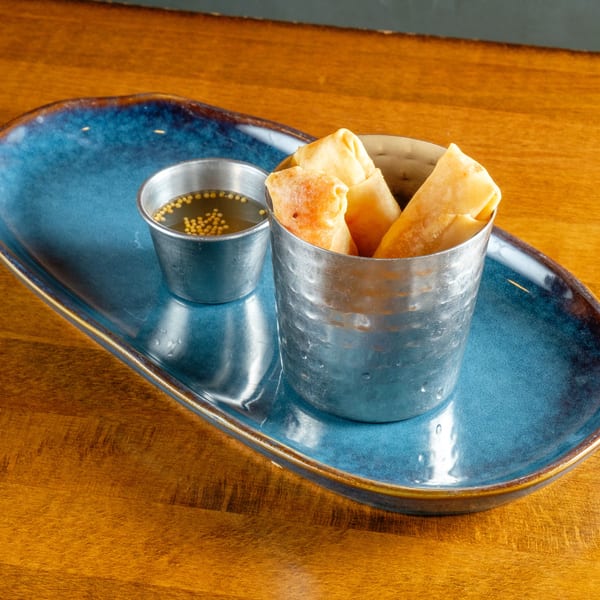 Crispy spring rolls in a metal cup with a side of dipping sauce on a blue plate.
