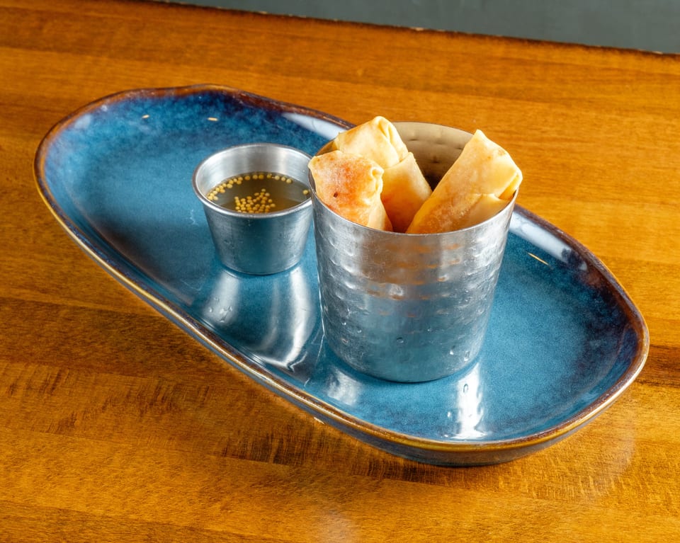 Crispy spring rolls in a metal cup with a side of dipping sauce on a blue plate.