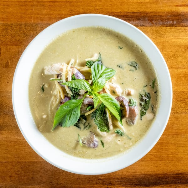 Overhead view of a bowl of green curry with chicken, bamboo shoots, and basil on a wooden table.