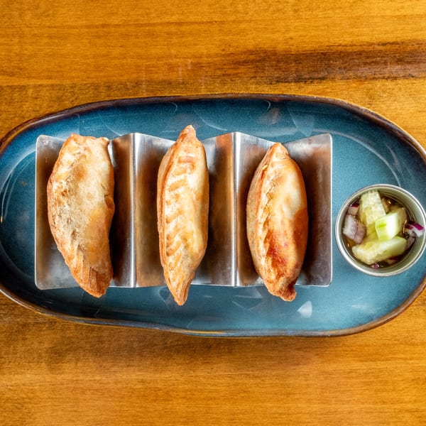 Three golden-brown empanadas in a metal holder on a blue plate with a side of pickled vegetables.