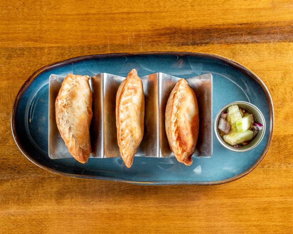 Three golden-brown empanadas in a metal holder on a blue plate with a side of pickled vegetables.