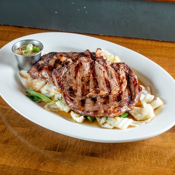 Grilled steak with grill marks, served on a bed of cabbage and green onions with a side of sauce.
