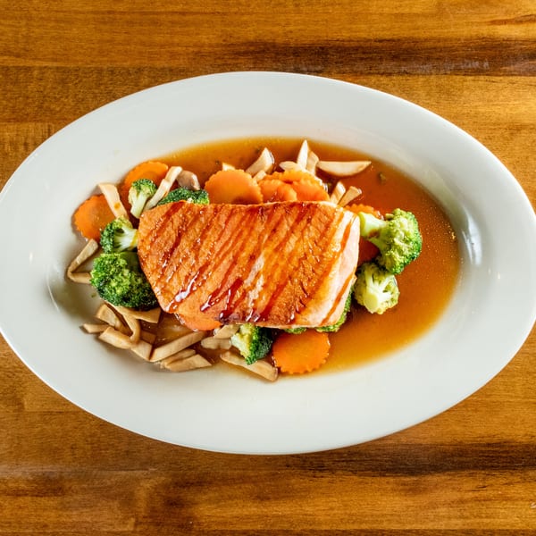 Grilled salmon fillet with broccoli, carrots, and mushrooms in a savory sauce on a white plate.