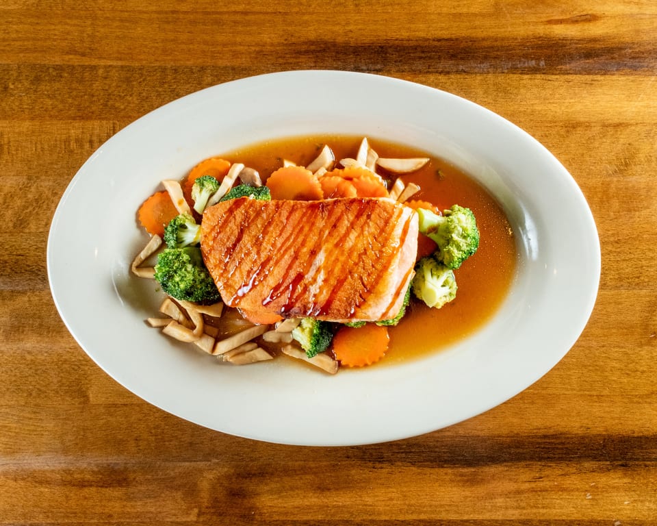 Grilled salmon fillet with broccoli, carrots, and mushrooms in a savory sauce on a white plate.