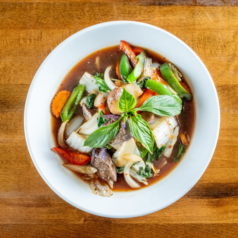 Overhead view of a colorful vegetable stir-fry with basil in a white bowl on a wooden table.