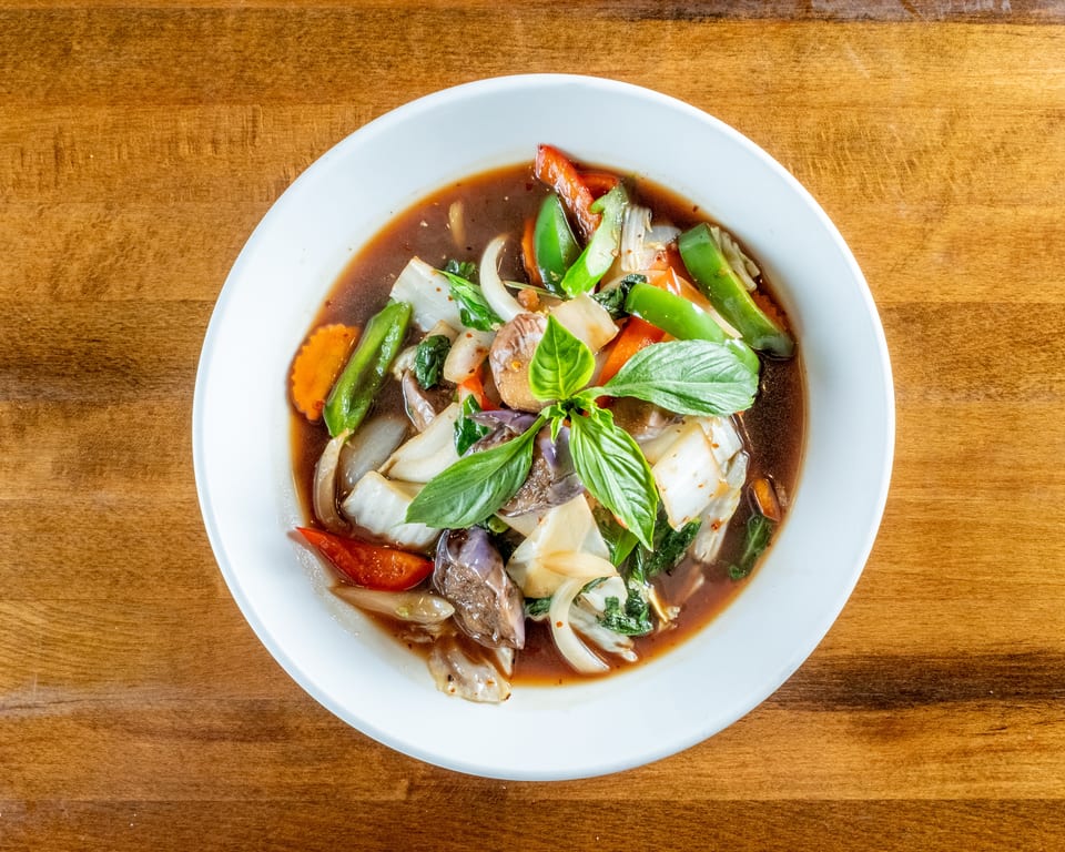 Overhead view of a colorful vegetable stir-fry with basil in a white bowl on a wooden table.