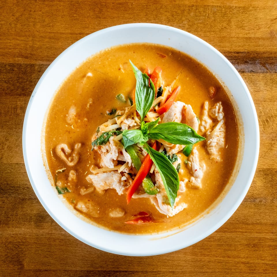 Overhead view of a white bowl filled with red curry, chicken, bamboo shoots, peppers, and basil.