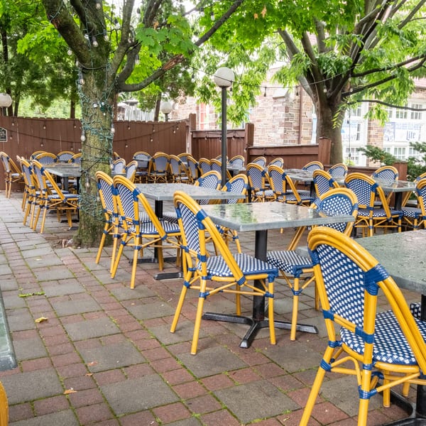 Outdoor patio dining area with many empty tables and blue and yellow woven chairs under green trees.
