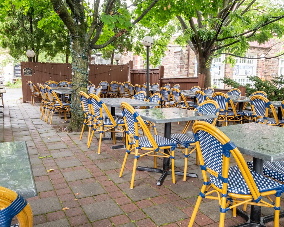 Outdoor patio dining area with many empty tables and blue and yellow woven chairs under green trees.