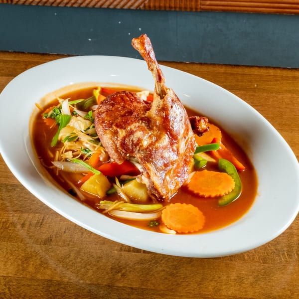 Crispy duck leg with vegetables in a rich brown sauce, served in a white oval bowl.