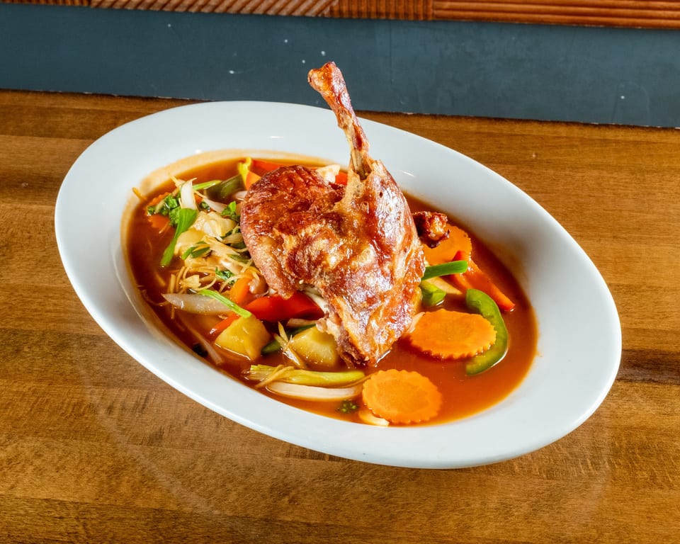 Crispy duck leg with vegetables in a rich brown sauce, served in a white oval bowl.