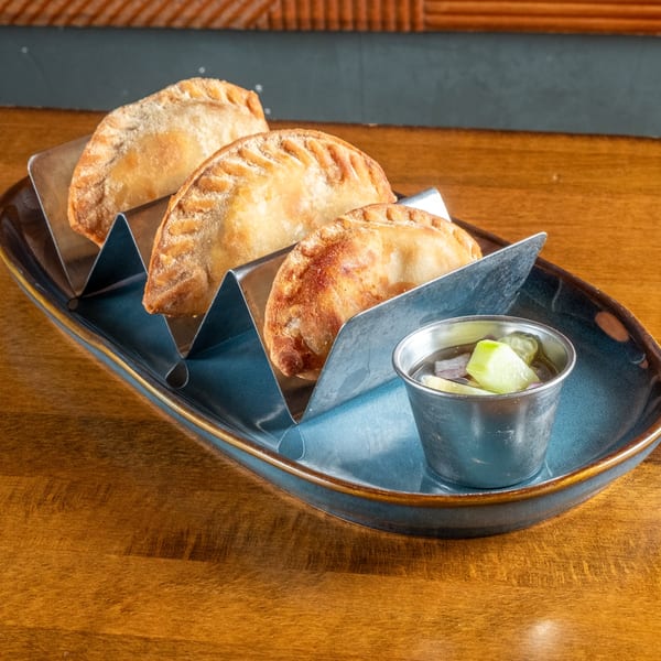 Three golden-brown empanadas in a metal holder on a blue plate with a side of dipping sauce.