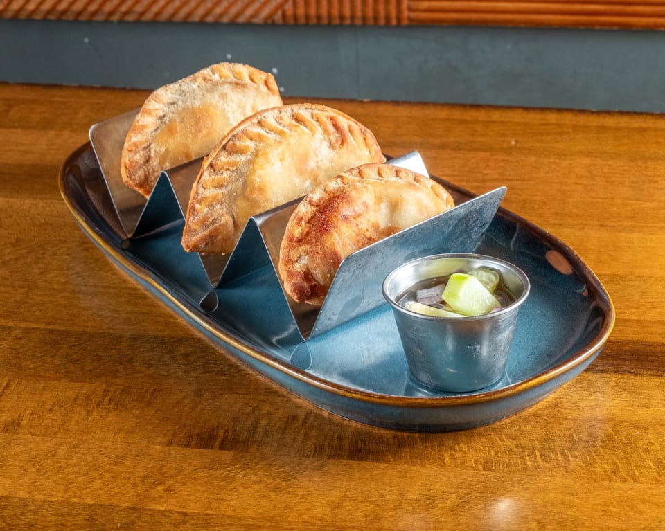 Three golden-brown empanadas in a metal holder on a blue plate with a side of dipping sauce.