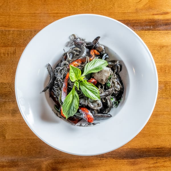 Squid ink pasta with seafood, red peppers, and basil in a white bowl on a wooden table.