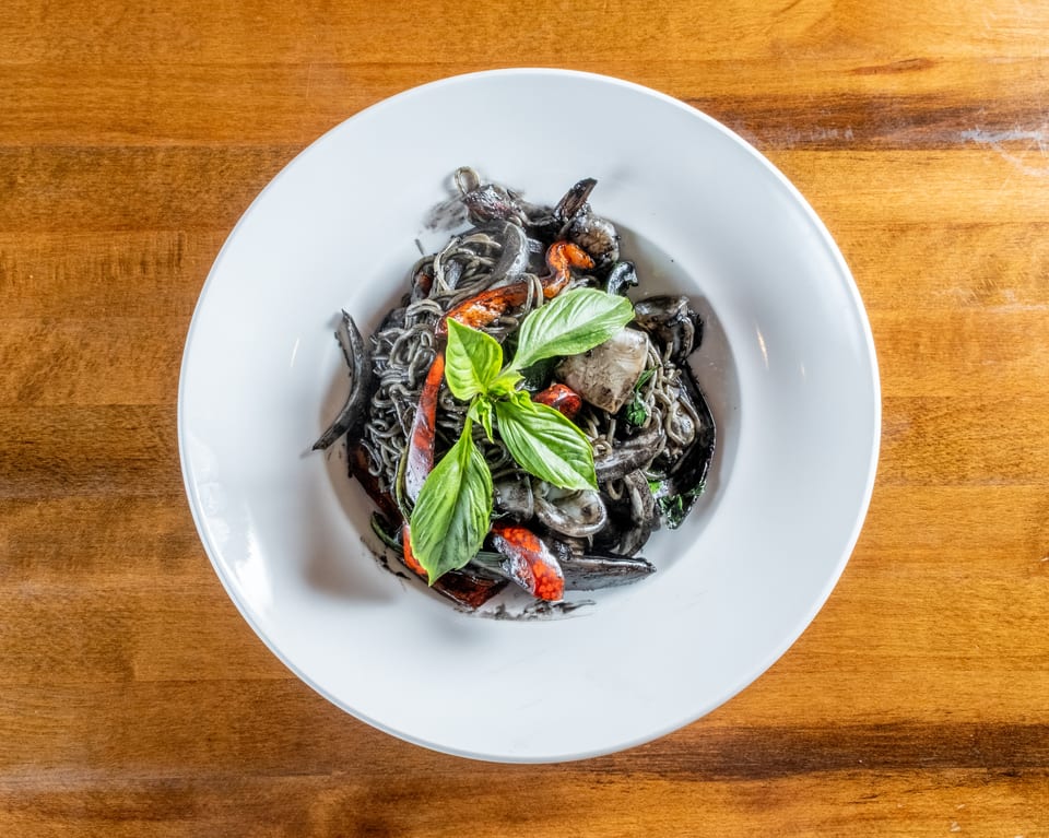 Squid ink pasta with seafood, red peppers, and basil in a white bowl on a wooden table.