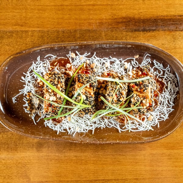 Four sesame-crusted tofu pieces with peanuts, scallions, and sauce on a bed of crispy noodles.