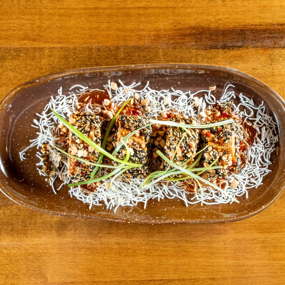 Four sesame-crusted tofu pieces with peanuts, scallions, and sauce on a bed of crispy noodles.