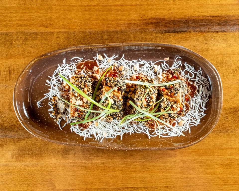 Four sesame-crusted tofu pieces with peanuts, scallions, and sauce on a bed of crispy noodles.