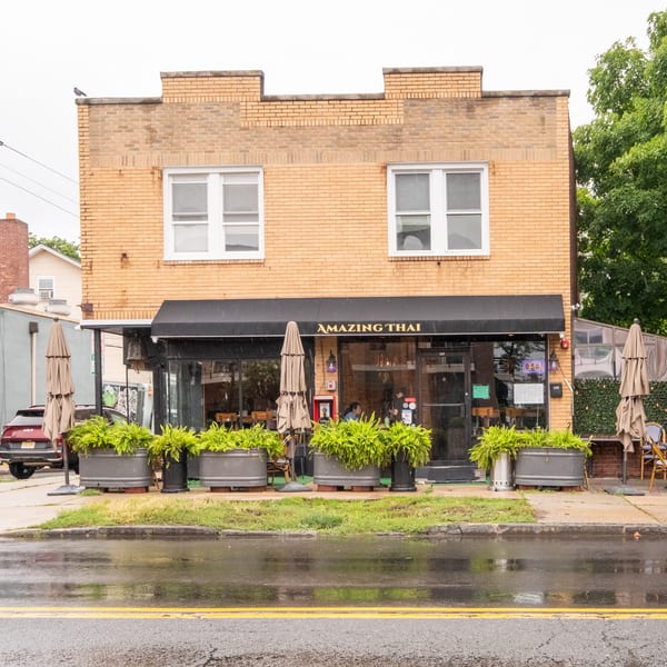 Amazing Thai restaurant exterior with a black awning, outdoor seating, and potted plants on a wet street.
