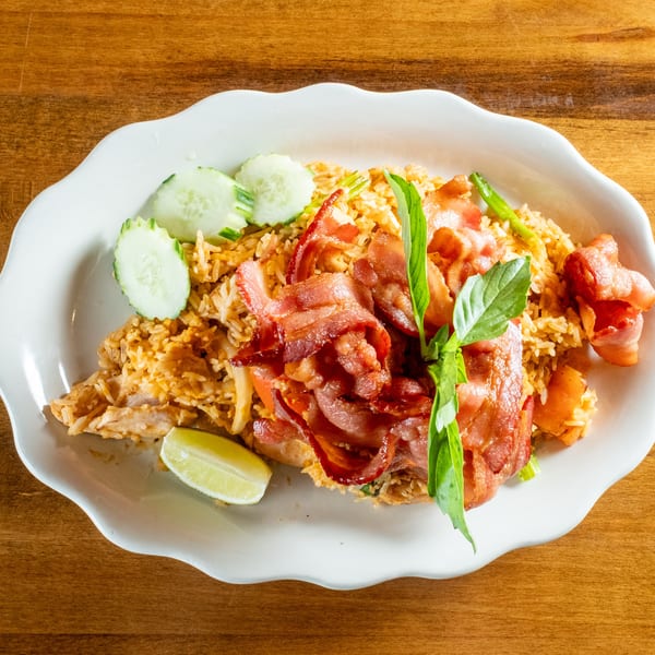 Fried rice with crispy bacon, fresh basil, cucumber slices, and a lime wedge on a white plate.