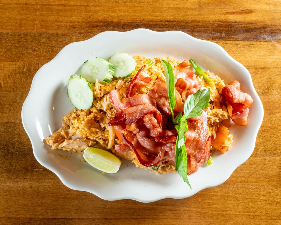 Fried rice with crispy bacon, fresh basil, cucumber slices, and a lime wedge on a white plate.