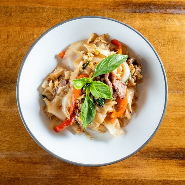 Pad See Ew with wide rice noodles, beef, bell peppers, and basil in a white bowl.