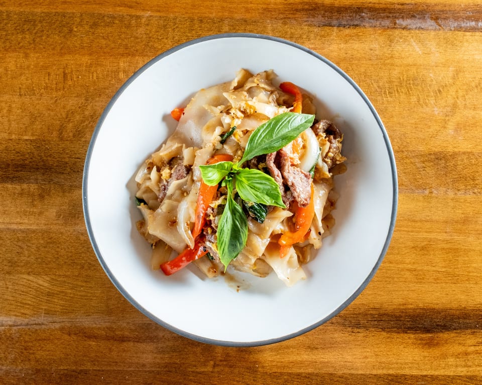 Pad See Ew with wide rice noodles, beef, bell peppers, and basil in a white bowl.