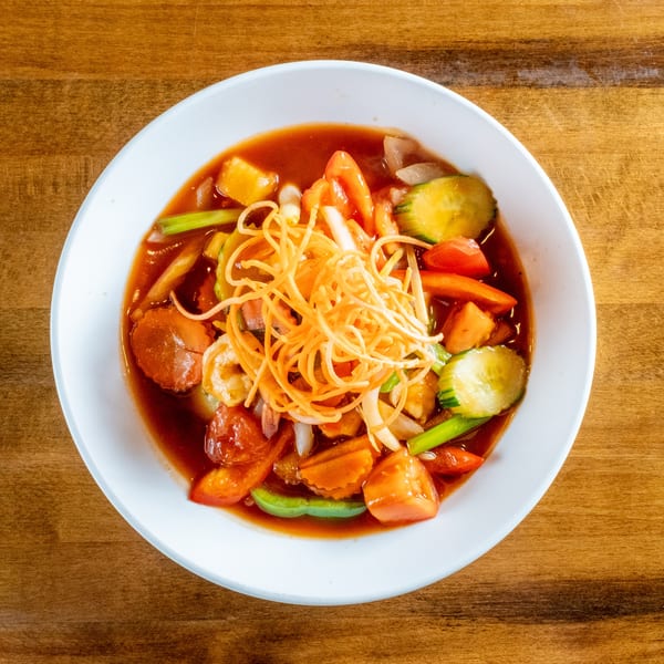Sweet and sour dish with shrimp, vegetables, and crispy noodles in a white bowl on wood.