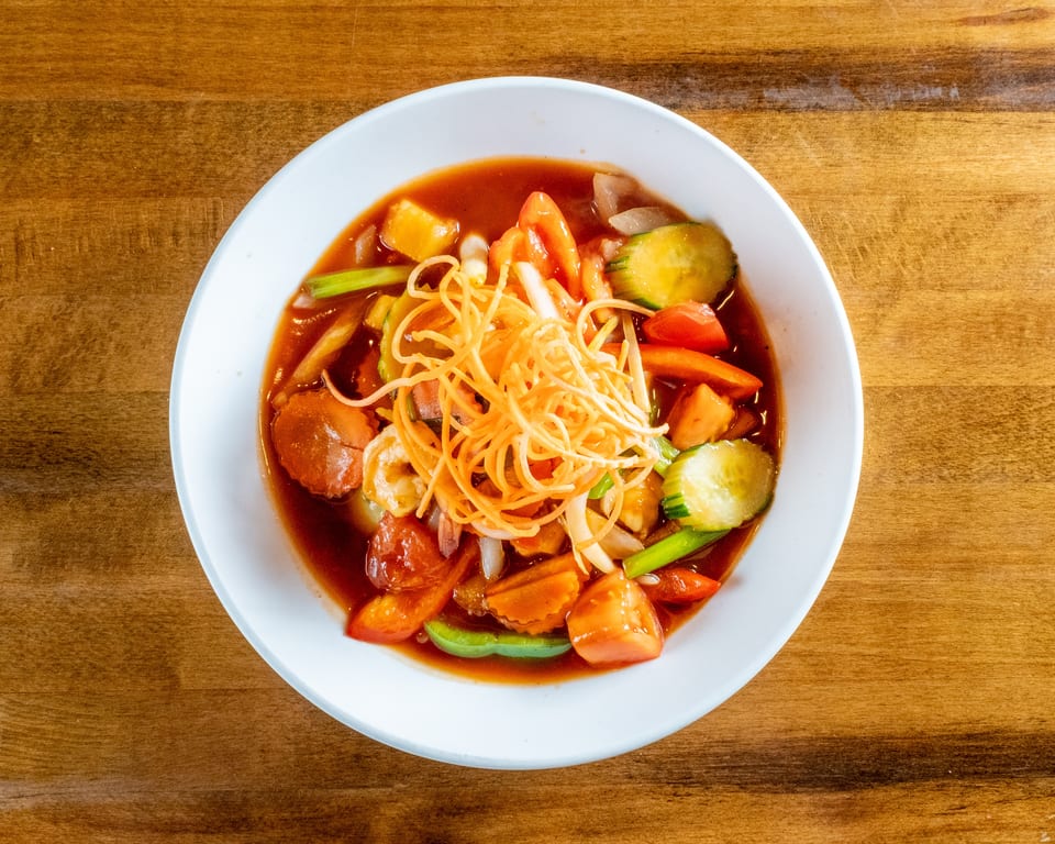 Sweet and sour dish with shrimp, vegetables, and crispy noodles in a white bowl on wood.