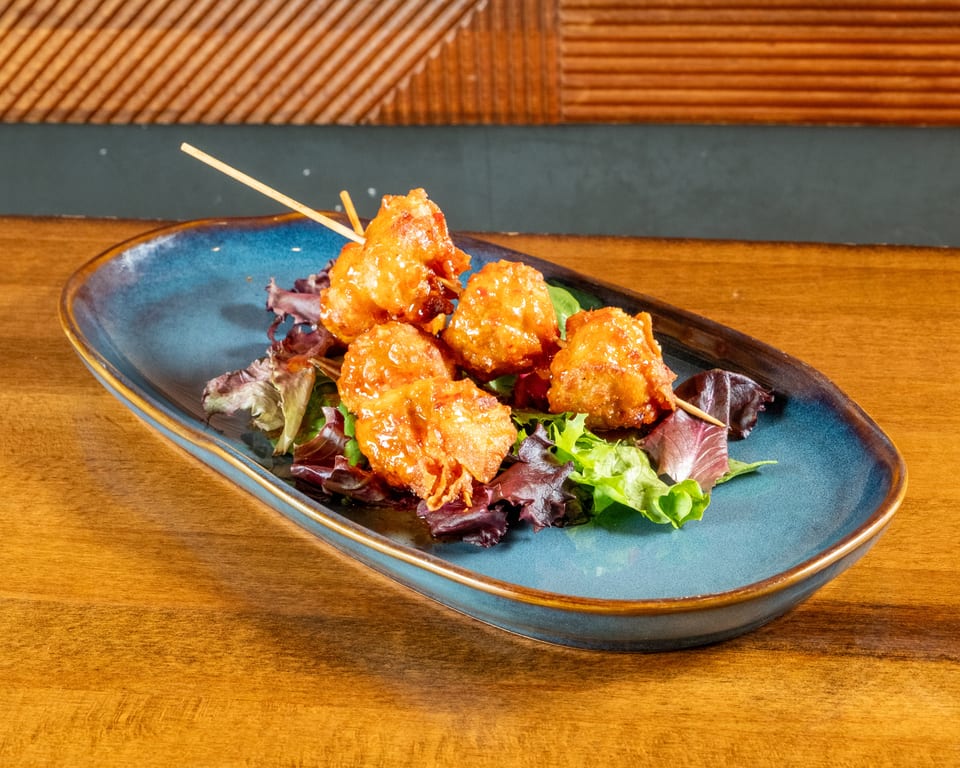 Five crispy, glazed chicken bites on skewers, served on a bed of mixed greens on a blue plate.