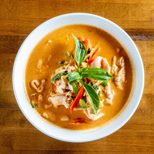 Red curry with chicken, bamboo shoots, bell peppers, and basil in a white bowl on a wooden table.