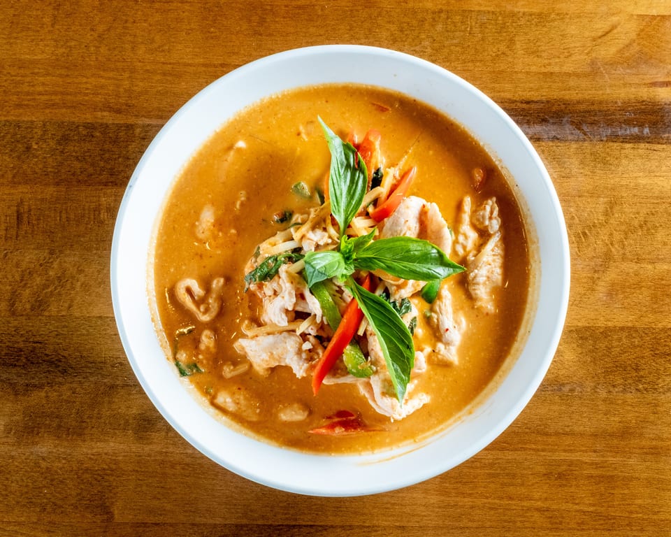 Red curry with chicken, bamboo shoots, bell peppers, and basil in a white bowl on a wooden table.