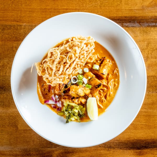 Overhead view of a bowl of Khao Soi with chicken, crispy noodles, and garnishes on a wooden table.