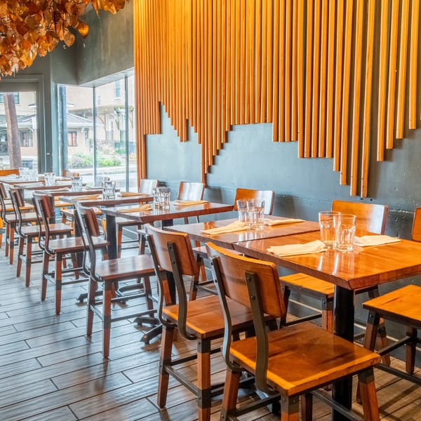 A modern restaurant interior with wooden tables and chairs, a decorative leaf ceiling, and a slatted wood wall.