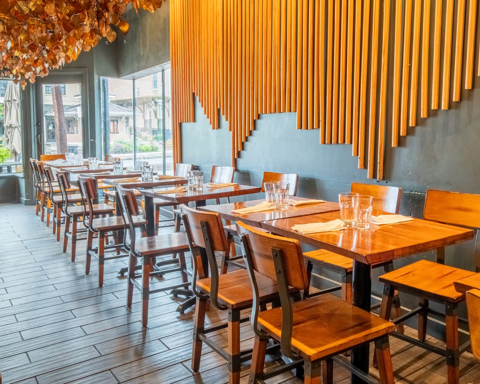 A modern restaurant interior with wooden tables and chairs, a decorative leaf ceiling, and a slatted wood wall.