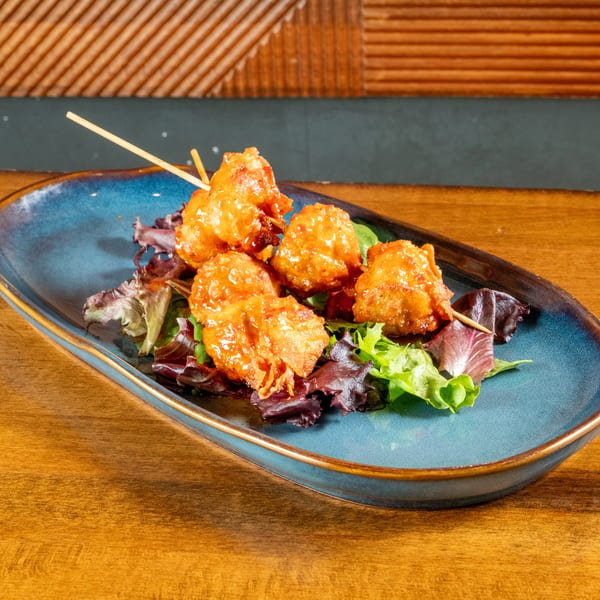 Five crispy, glazed chicken bites on skewers, served on a bed of mixed greens on a blue plate.