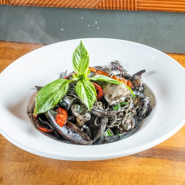 Steaming black squid ink pasta with seafood, tomatoes, and basil in a white bowl.