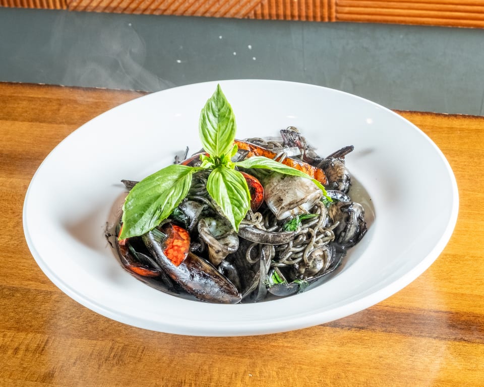 Steaming black squid ink pasta with seafood, tomatoes, and basil in a white bowl.