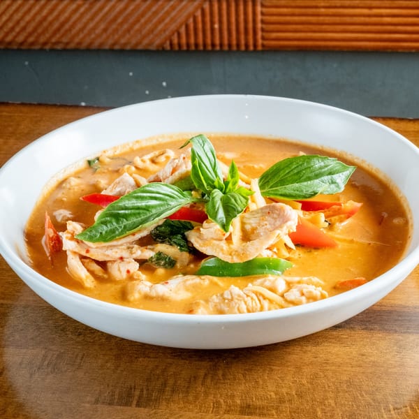 A white bowl of red curry with chicken, red and green peppers, and basil on a wooden table.