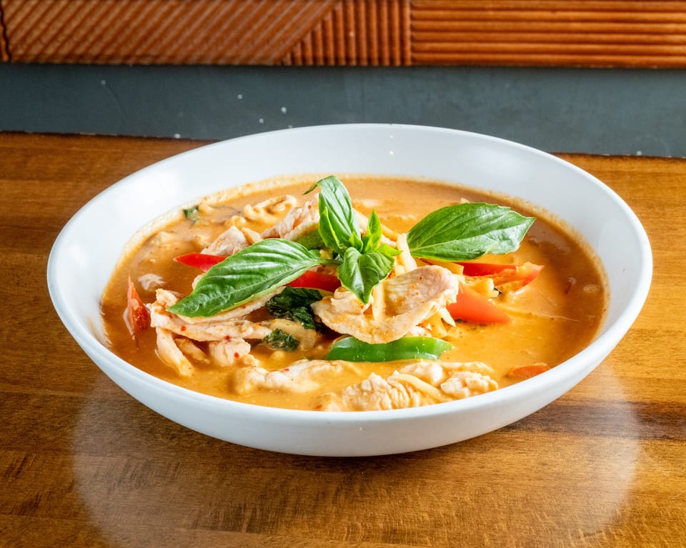A white bowl of red curry with chicken, red and green peppers, and basil on a wooden table.