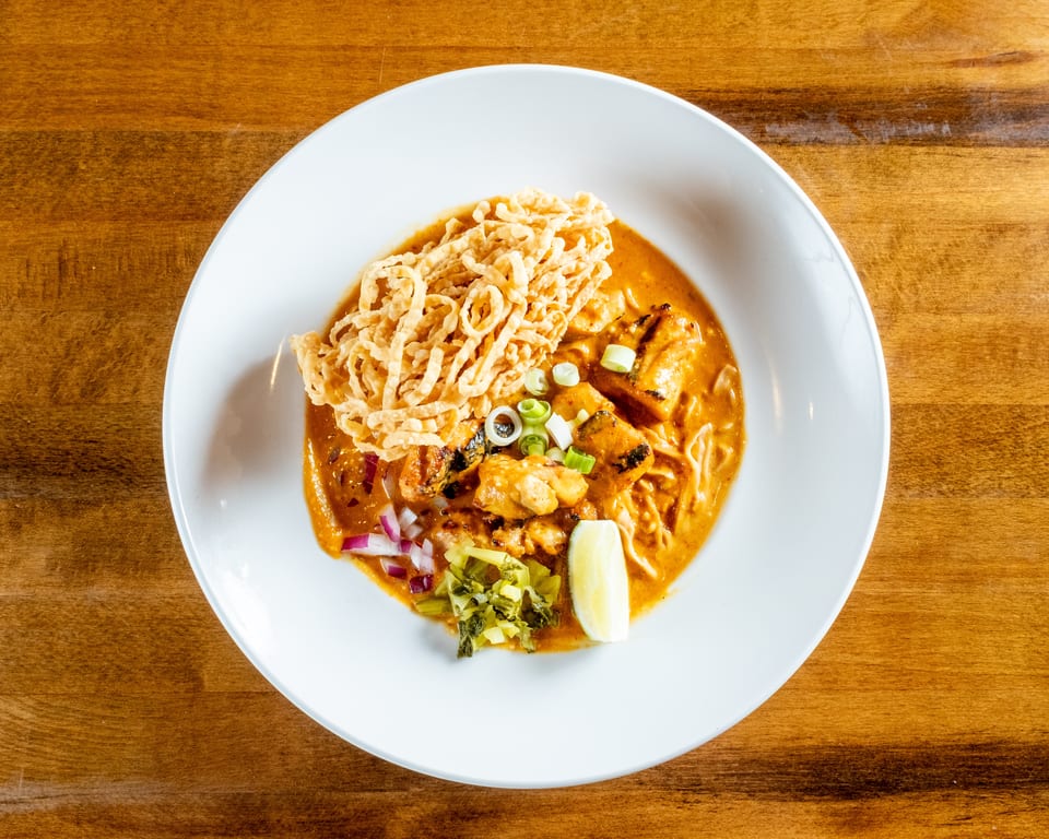 Overhead view of a bowl of Khao Soi with chicken, crispy noodles, lime, and red onion.