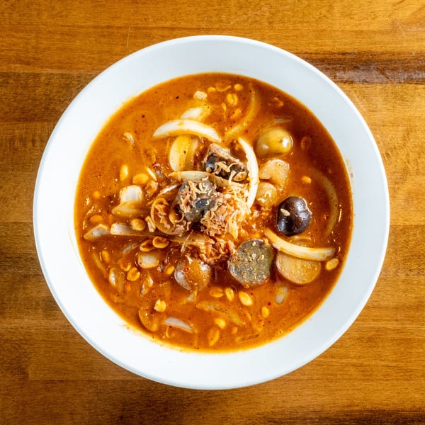 Overhead view of a bowl of spicy soup with onions, peanuts, and dark mushrooms on a wooden table.