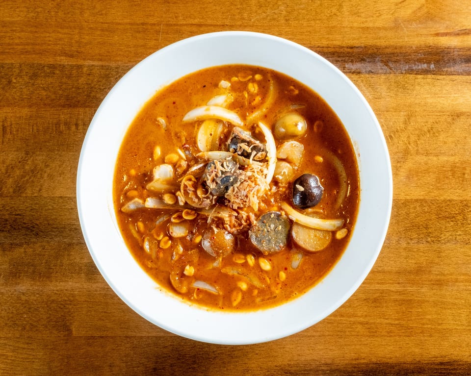 Overhead view of a bowl of spicy soup with onions, peanuts, and dark mushrooms on a wooden table.